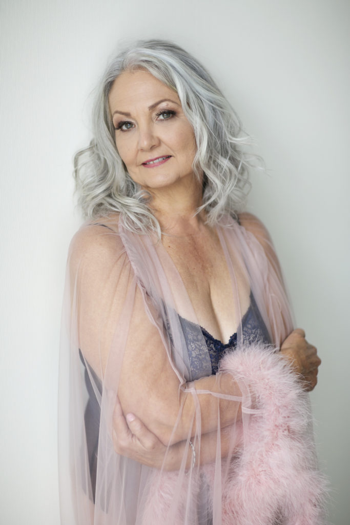 woman over 50 with pick fur robe smiling at camera during Boudoir session against a white wall